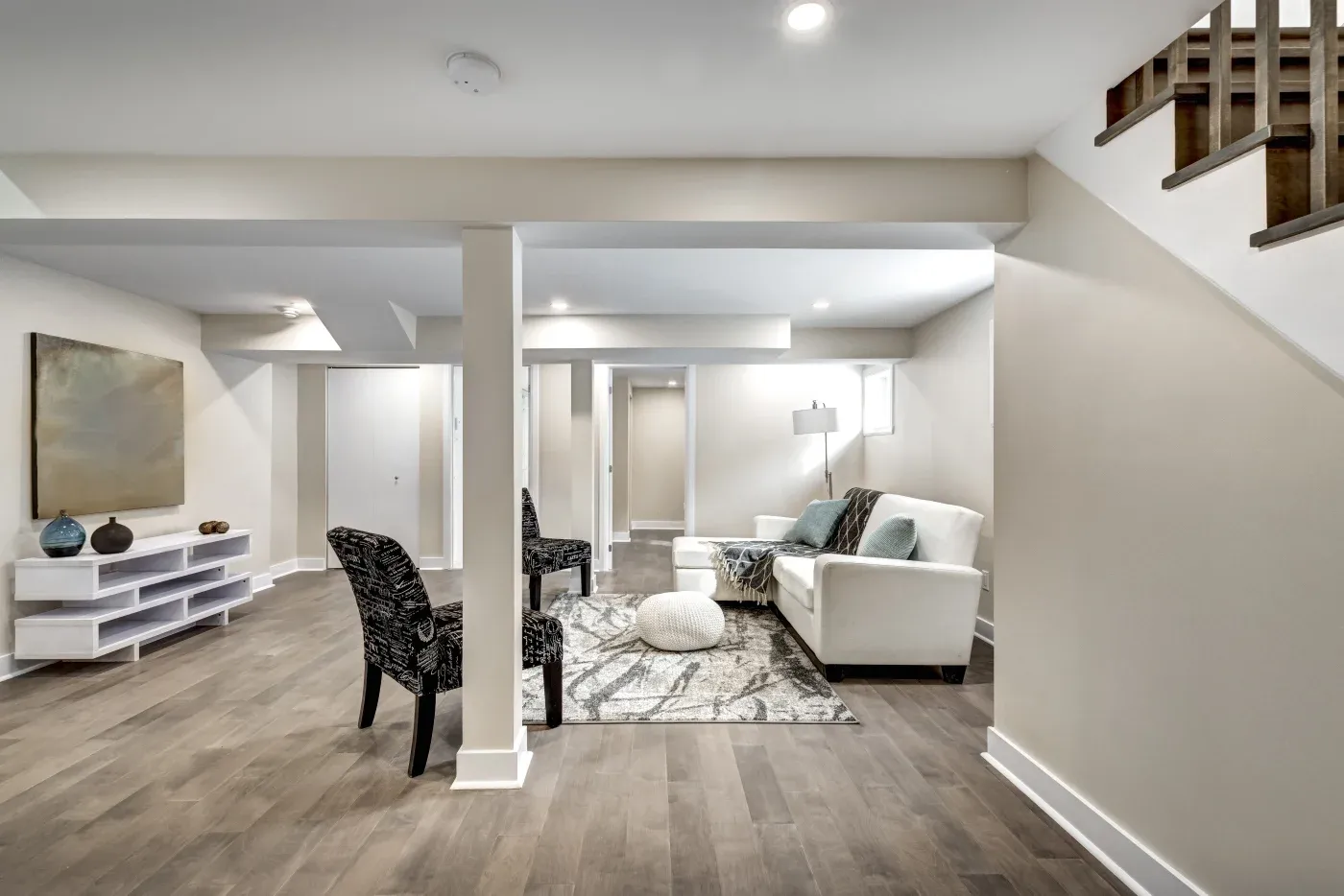 A modern, spacious basement with beige walls and wood flooring. It features a white sofa, patterned chairs, a gray rug, and wall art, creating a cozy ambiance.