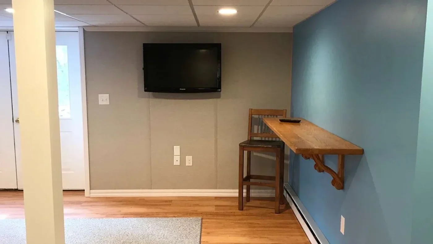 A minimalist room featuring a wall-mounted TV, a blue accent wall, a wooden bar with a tall chair, light wood flooring, and cozy lighting.