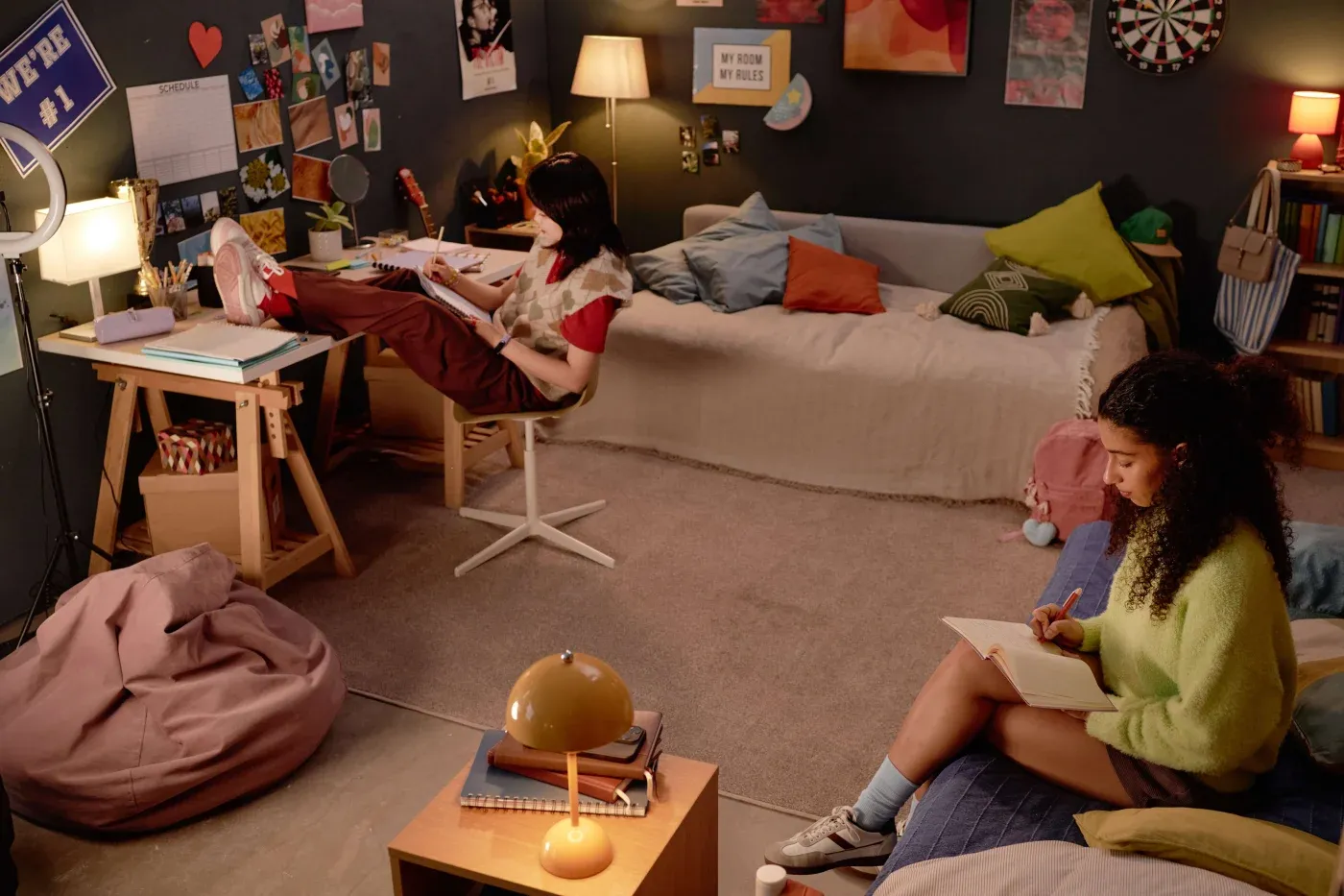 Teen bedroom with two girls studying. One is on a chair with feet on a desk, surrounded by books and posters. The other sits on a bed, writing. Cozy and focused atmosphere.