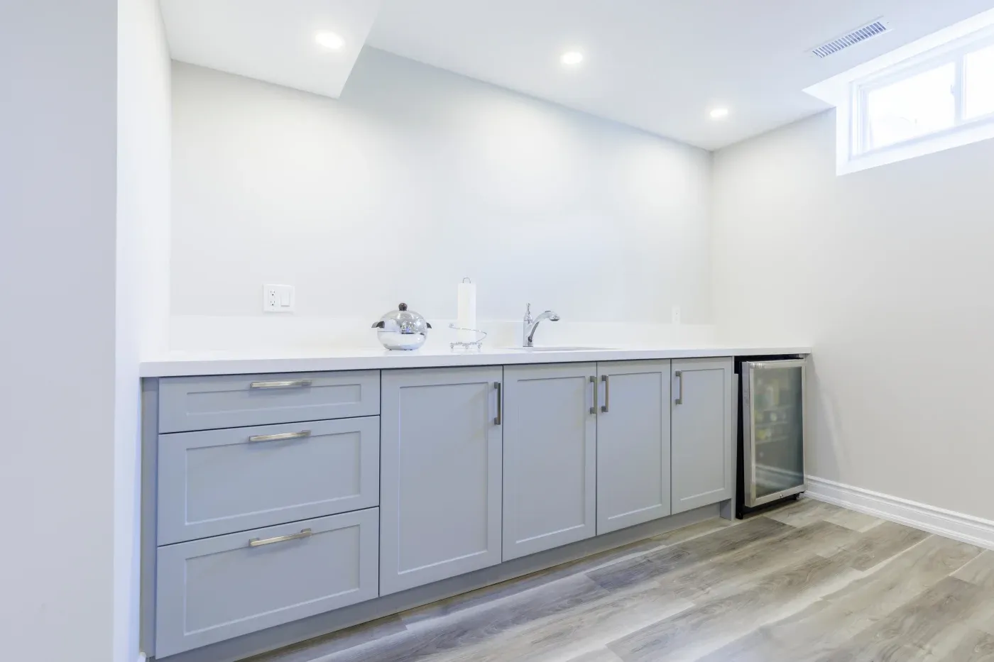 Modern kitchen with grey cabinets, white countertop, and a small silver fridge. Bright lighting and minimal decor create a clean, spacious feel.