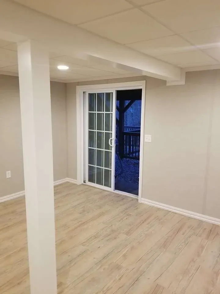 A freshly renovated basement room with light wood flooring, beige walls, and a sliding glass door leading outside. The space appears bright and inviting.