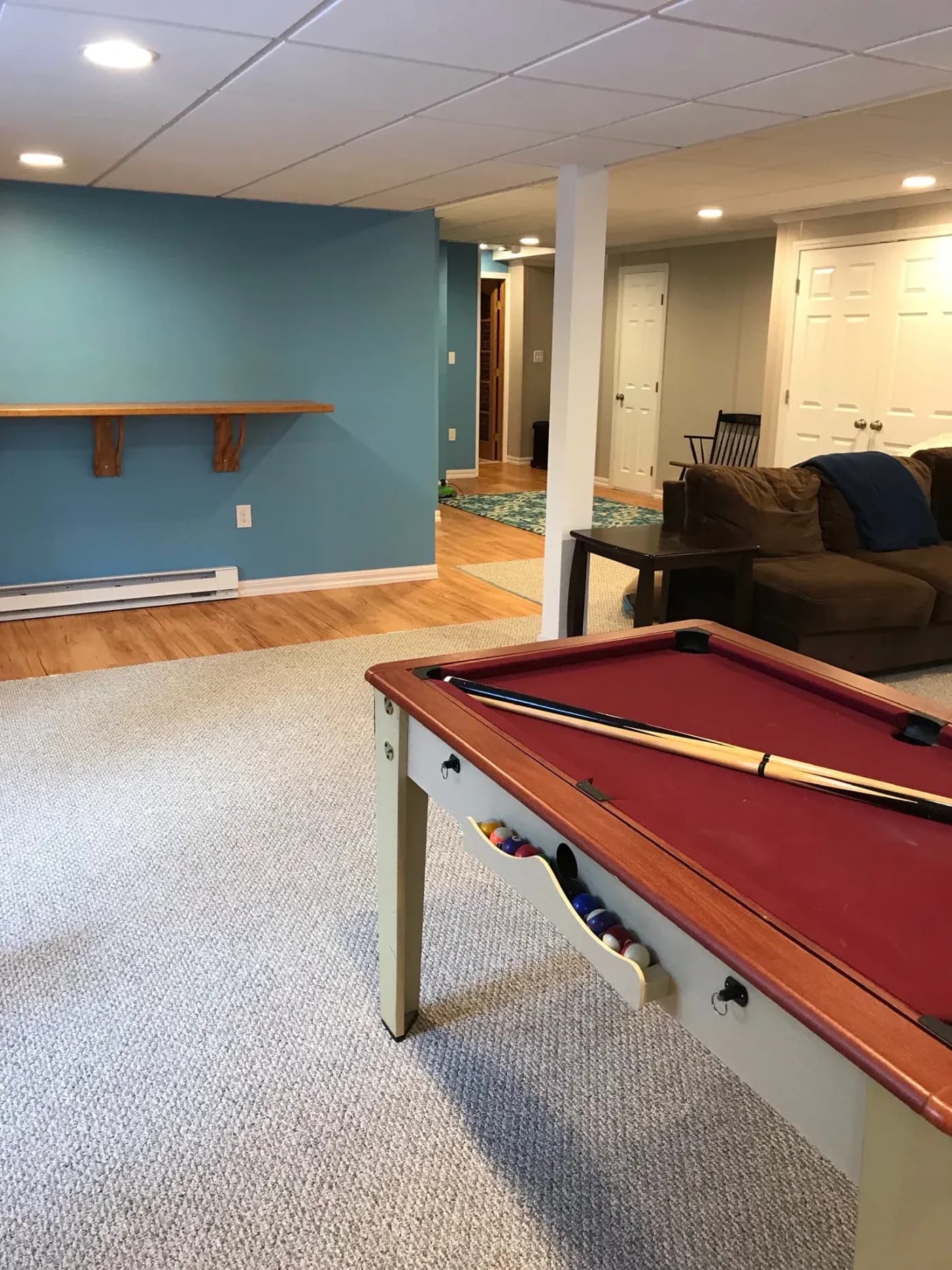 A cozy basement game room with a red pool table, blue accent wall, and a brown couch. The area has a relaxed atmosphere with warm lighting.