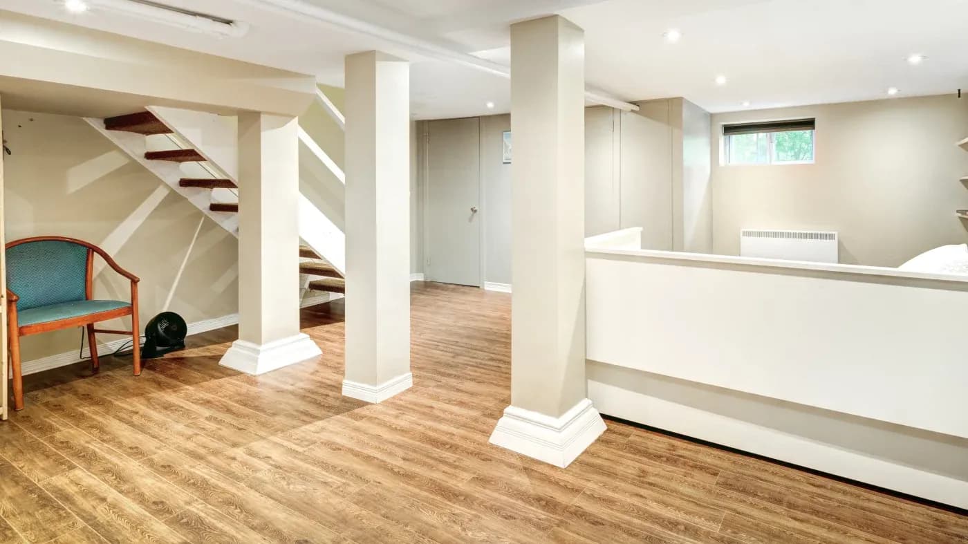 Spacious basement with wooden floors, white walls, and support columns. A staircase is on the left, next to a green chair, creating a clean, modern feel.