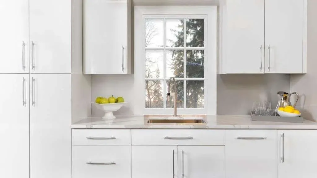 Modern kitchen with glossy white cabinets and drawers. A window with a view of trees above the sink. Green apples and a tray with lemons add freshness.