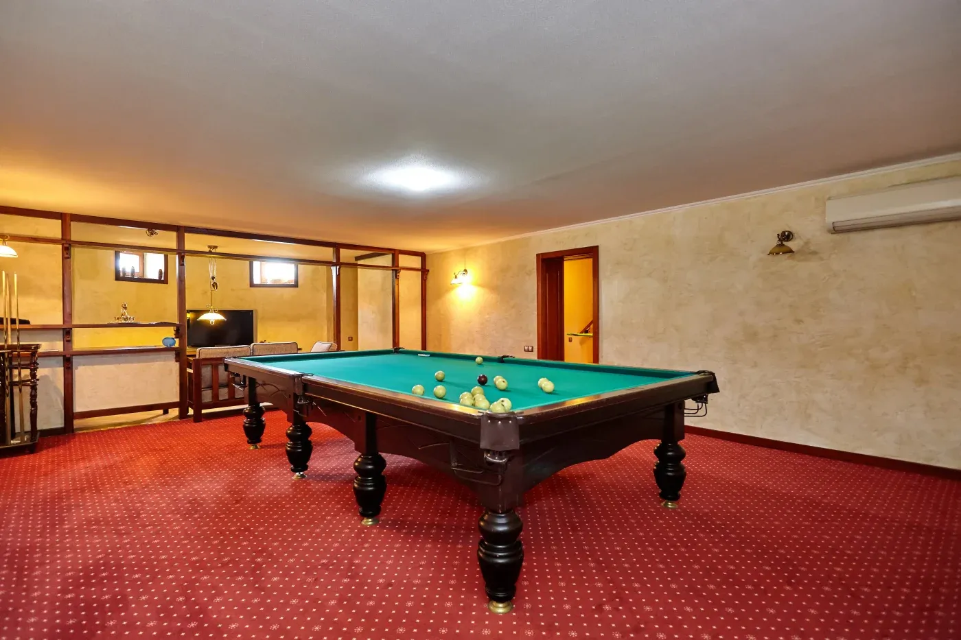 A spacious billiard room with a green pool table covered in balls, set on a red carpeted floor. The room has warm lighting and a television in the corner.