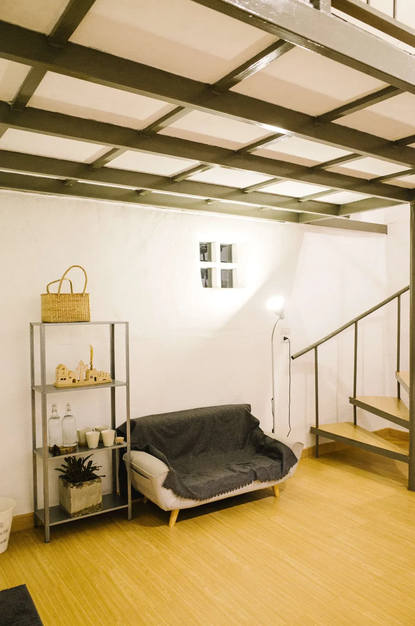A cozy, modern loft with wooden floors and a white wall, featuring a small couch, a standing lamp, metal stairs, and a shelf with decor items.