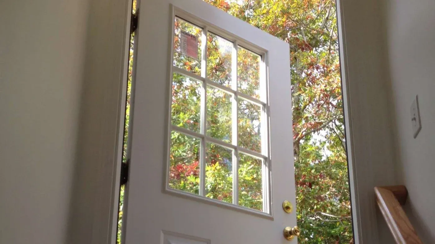 Open white door to vibrant autumn-colored trees outside. Sunlight filters through glass panels, creating a warm, inviting atmosphere.