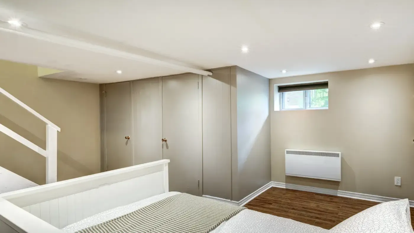A cozy basement bedroom with beige walls, recessed lighting, and a small window. A bed with a knitted blanket and a white heater complement the wood floor.