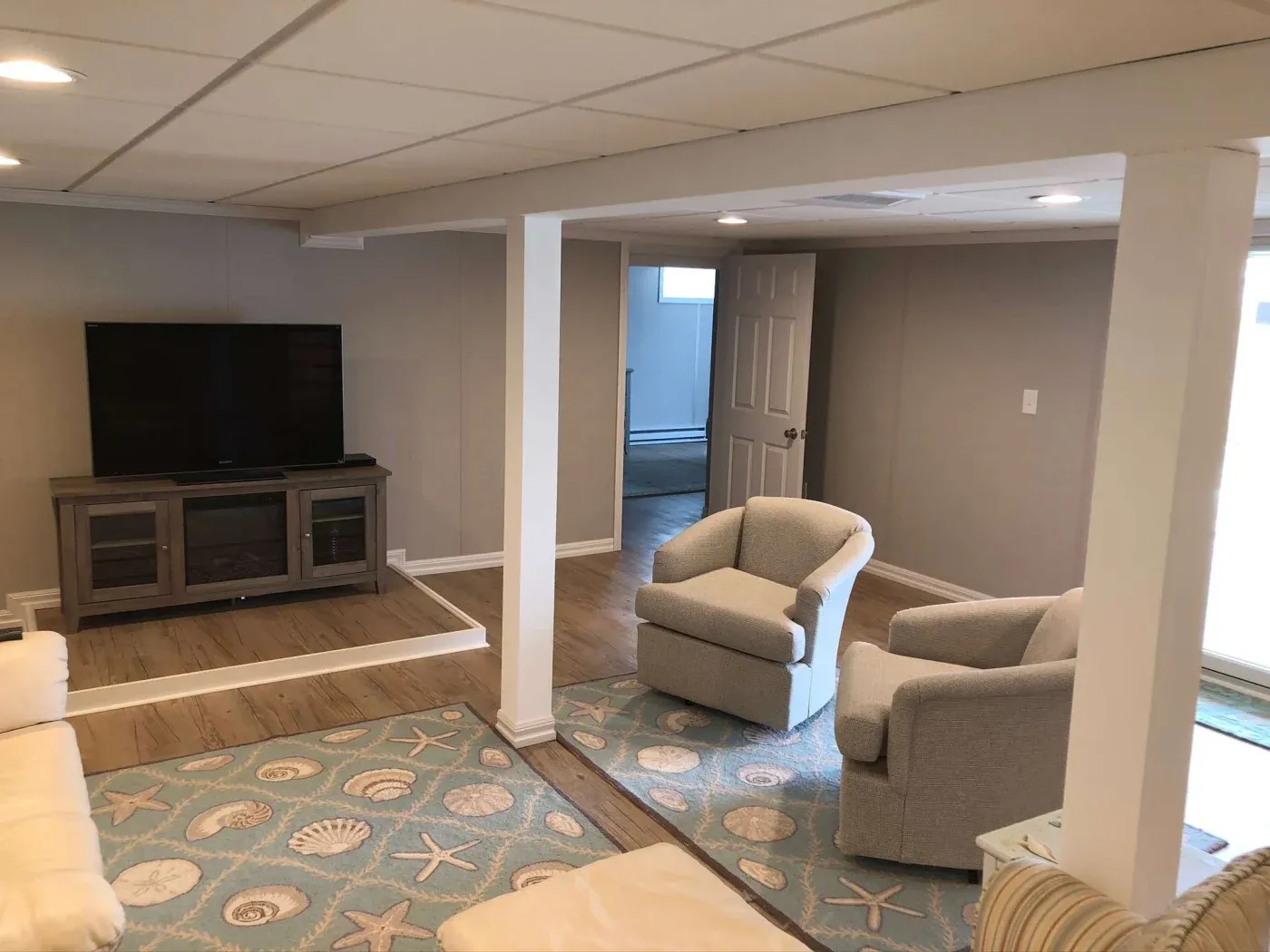 Cozy basement with soft lighting, featuring a TV on a stand, light beige chairs, and a coastal-themed rug on wooden floors. Relaxed atmosphere.