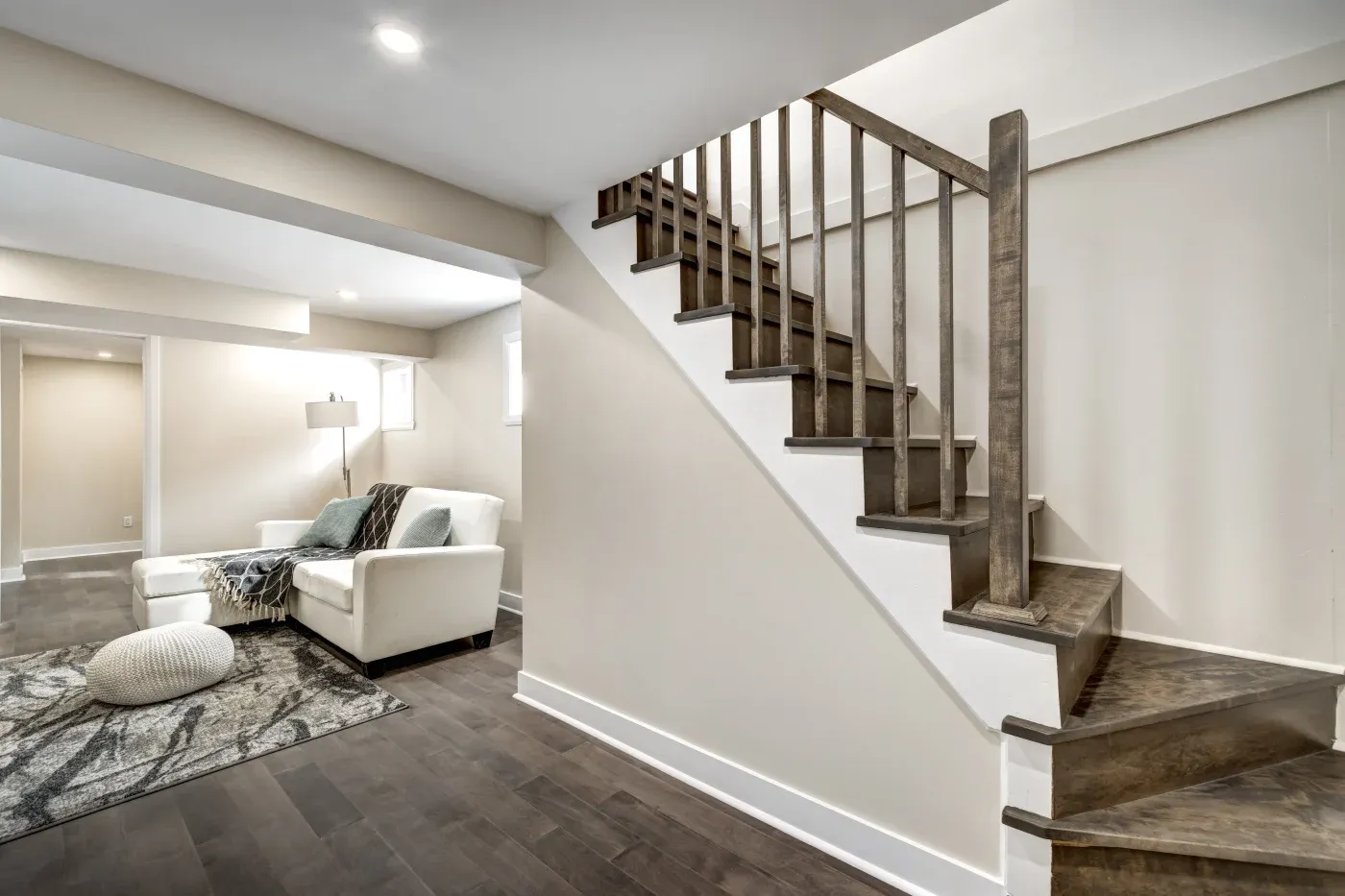 A modern basement with dark wood flooring features a staircase with wooden railings. A cozy corner includes a white armchair, a lamp, and a patterned rug.