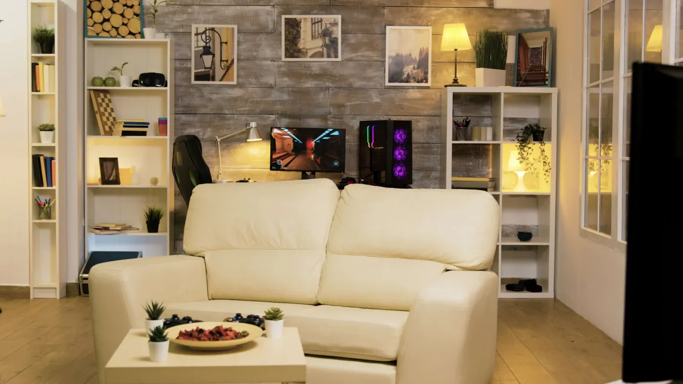 Cozy living room with a cream sofa, small potted plants, and a coffee table. In the background, shelves, art, and a lit computer setup on a desk.
