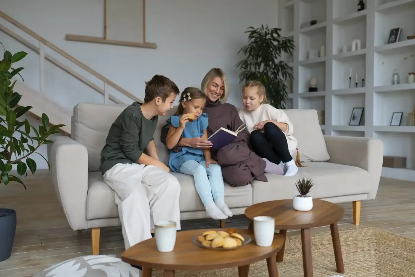 A family of four sits on a cozy beige sofa in a modern living room, reading a book together. The atmosphere is warm and relaxed, with cookies and drinks on the table.