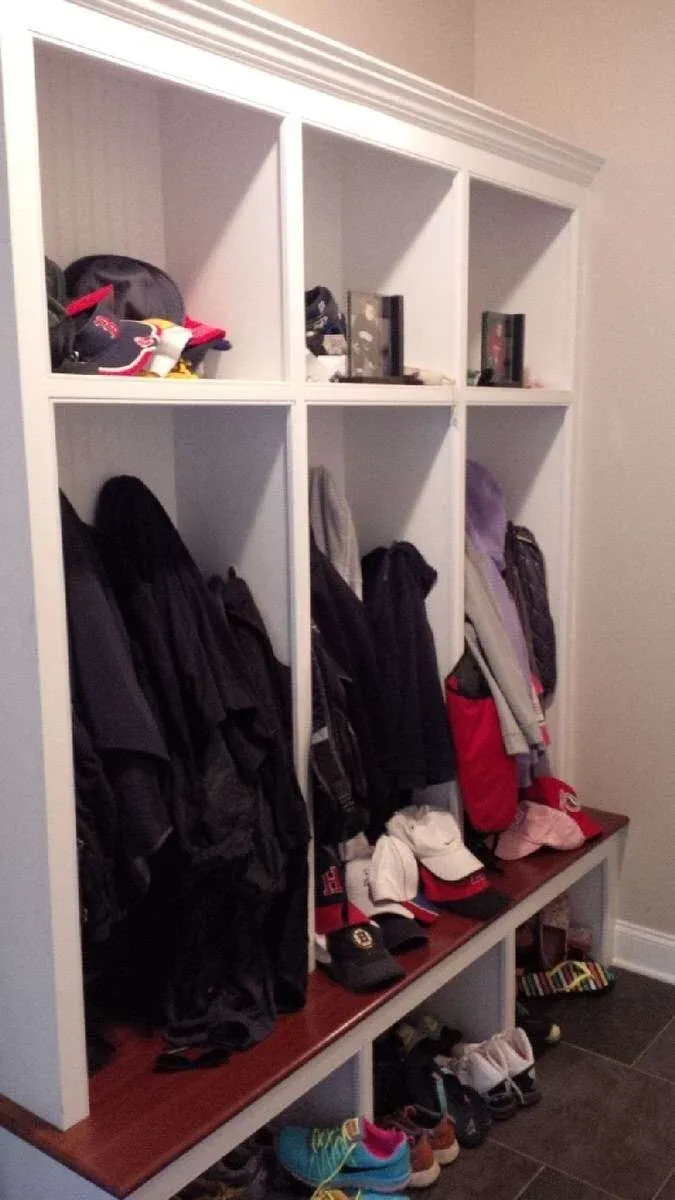 A white cubby unit with four sections, each holding coats, hats, and framed photos. Below, assorted shoes and sneakers are neatly arranged on a wooden bench.