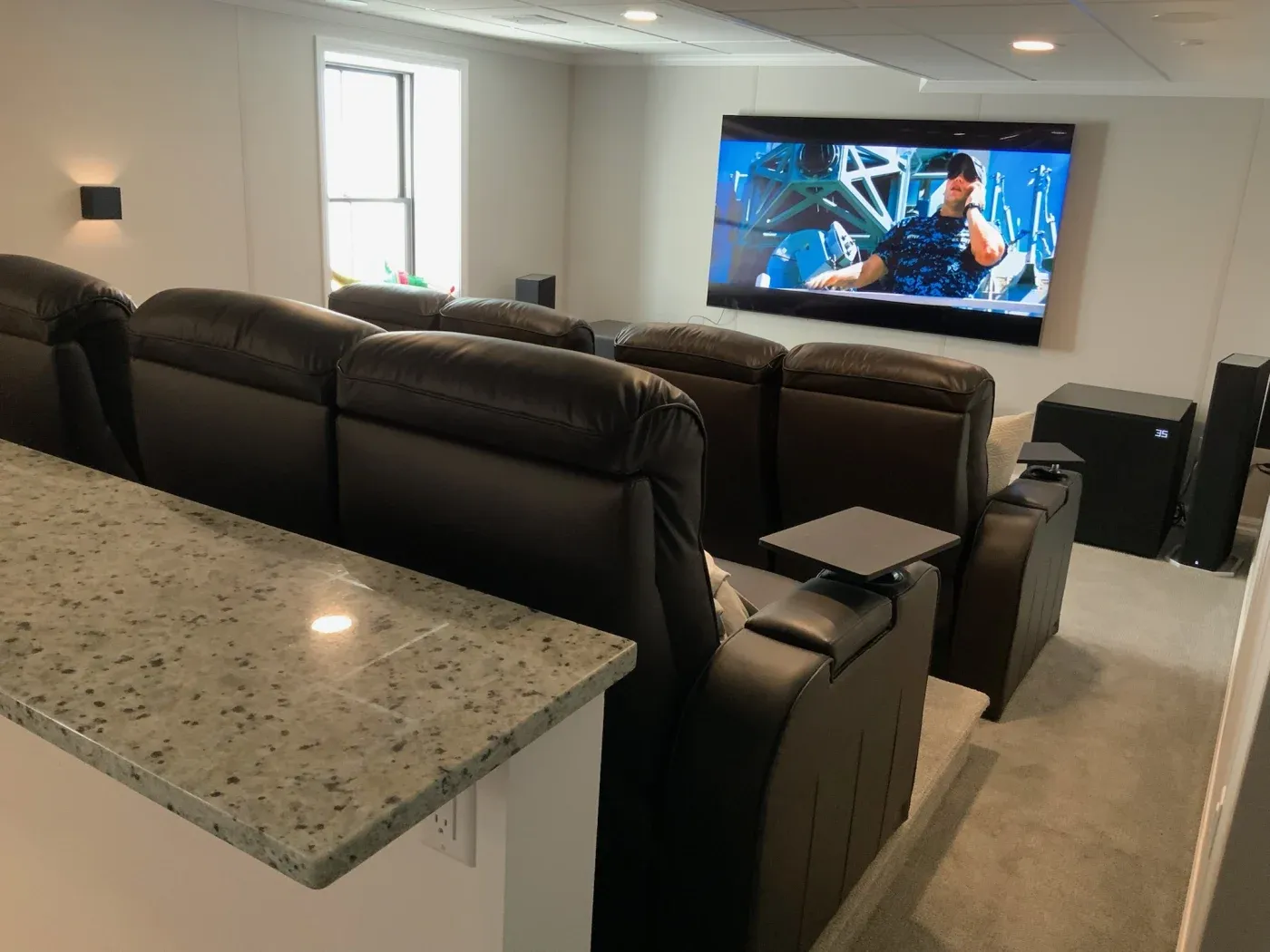 Home theater with black leather recliners facing a large screen displaying a movie scene. A granite countertop is in the foreground, evoking a cozy ambiance.