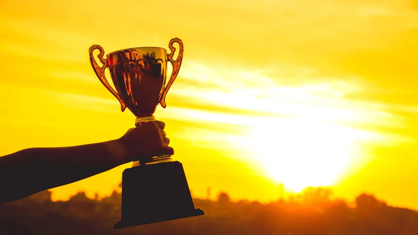 A hand holds up a golden trophy against a vibrant sunset, symbolizing victory and achievement. The background features silhouettes of trees under a glowing sky.