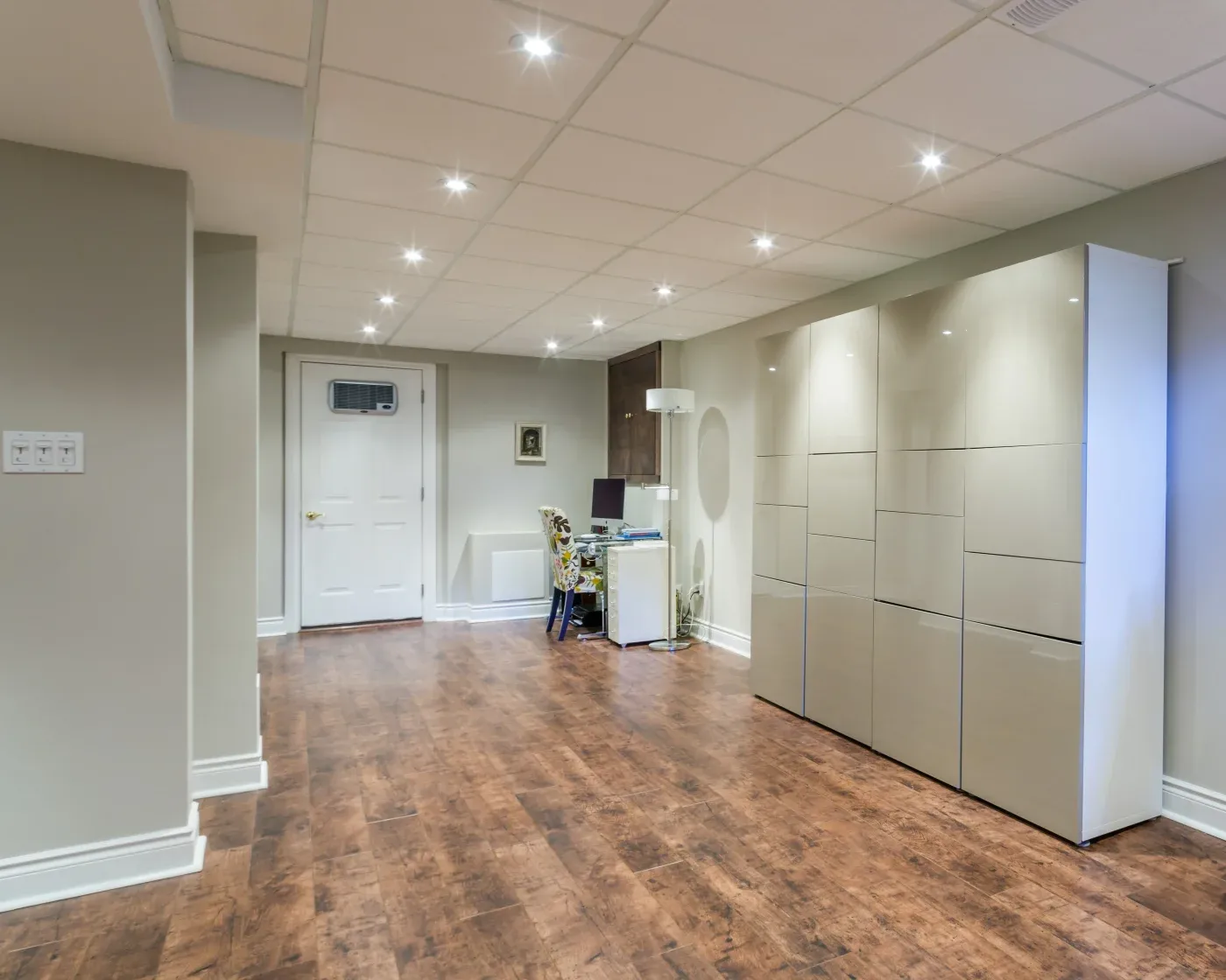 Spacious, modern basement with glossy storage cabinets, a small desk setup, and wood flooring. Bright, minimalist design with recessed lighting.