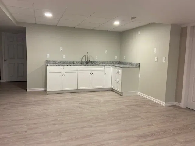 Minimalist modern kitchenette in a bright basement room with light wood flooring, white cabinets, marble countertop, and recessed lighting.