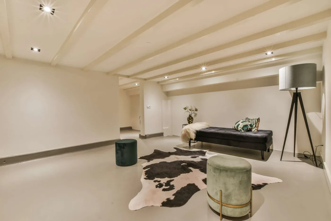 A minimalist room features a black daybed with a fur throw, green cushions, and a floor lamp. A cowhide rug lies on the floor between two plush, round stools. The beige walls and ceiling beams create a cozy, modern atmosphere.
