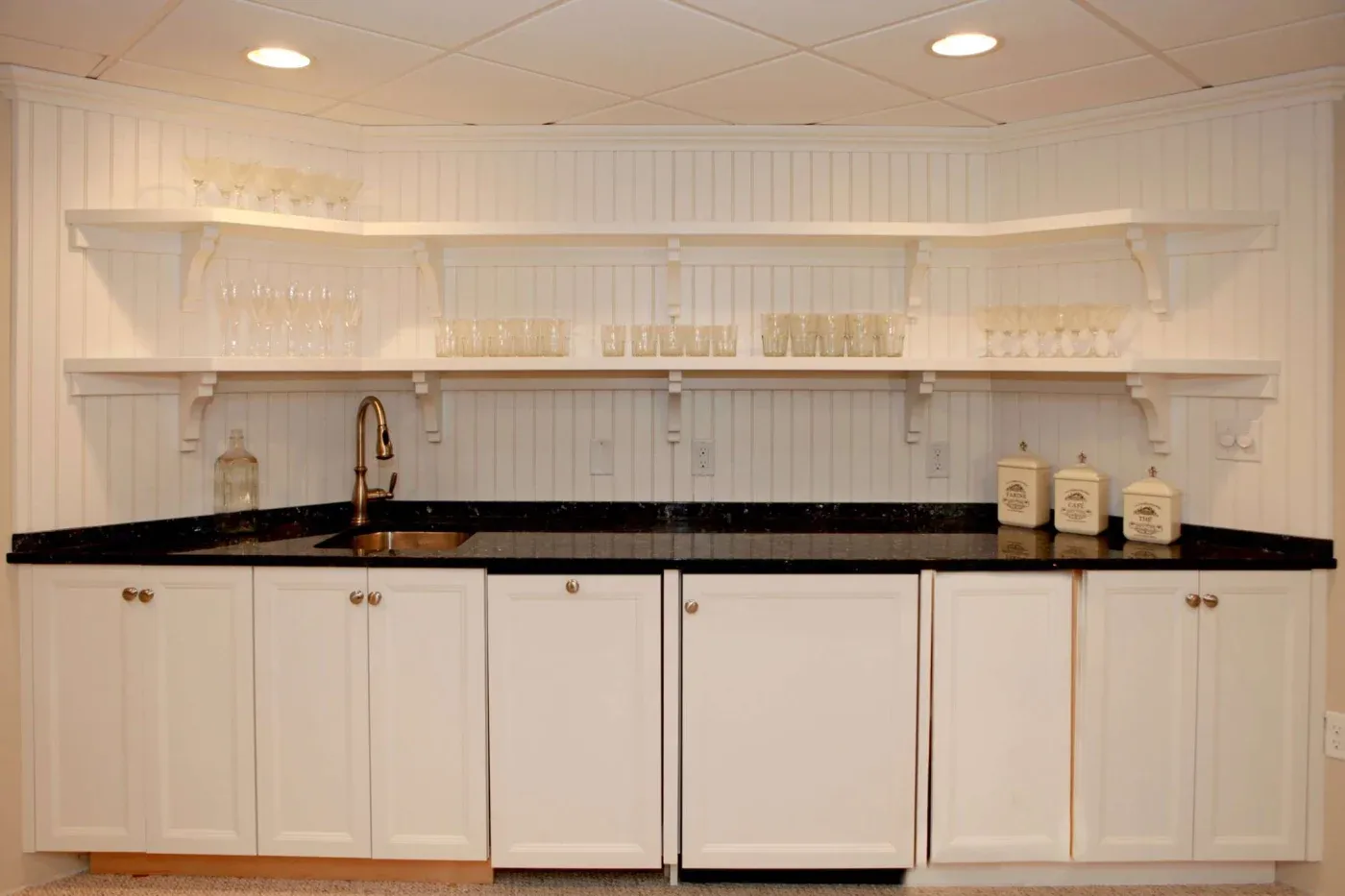 A home bar with white cabinets and black countertop, featuring open shelves with glassware. A small sink and decorative jars add a minimalist touch.