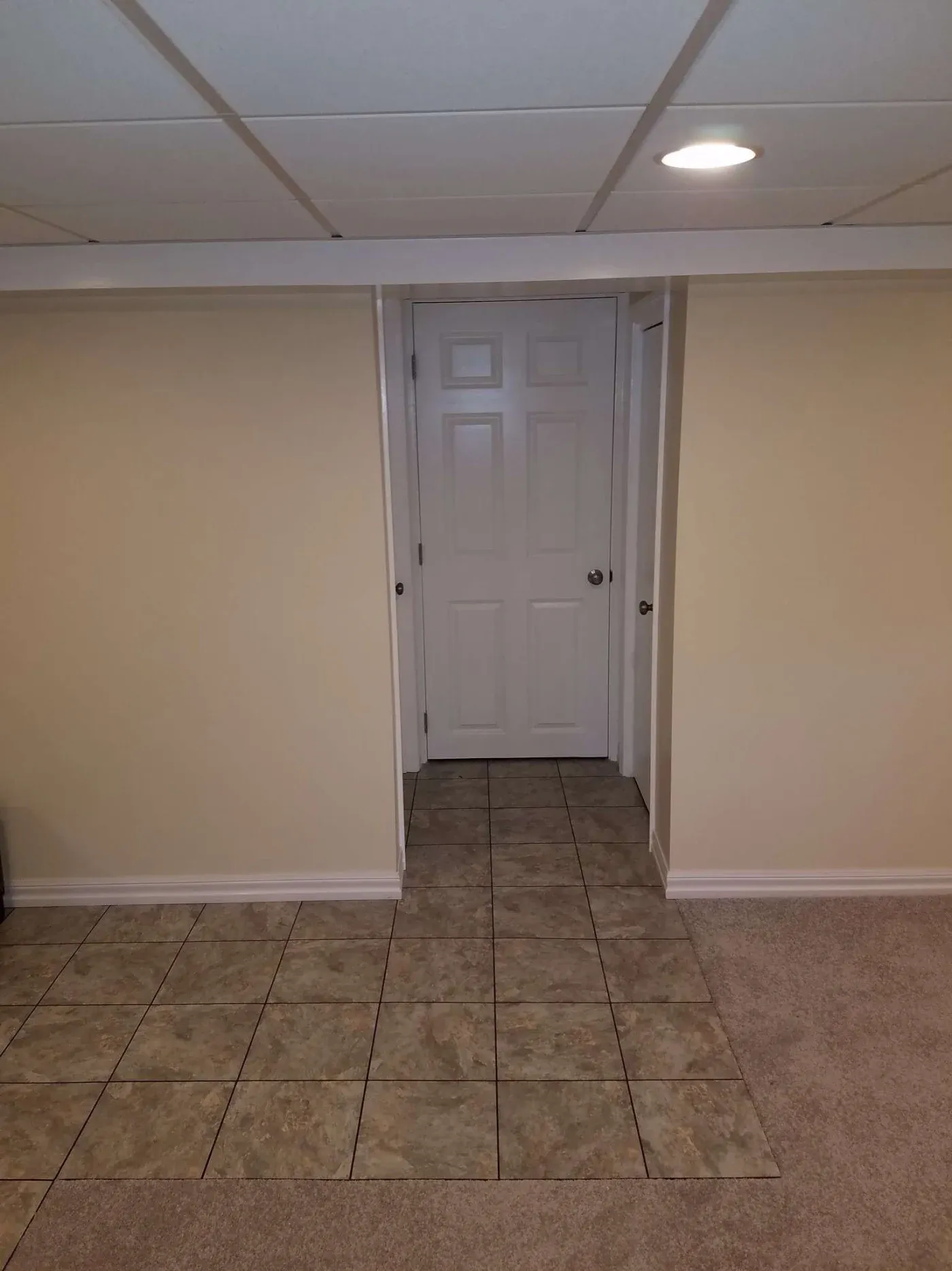 A small, narrow hallway with beige walls and a white door at the end. The floor is tiled in the hallway and carpeted in the foreground, under soft lighting.