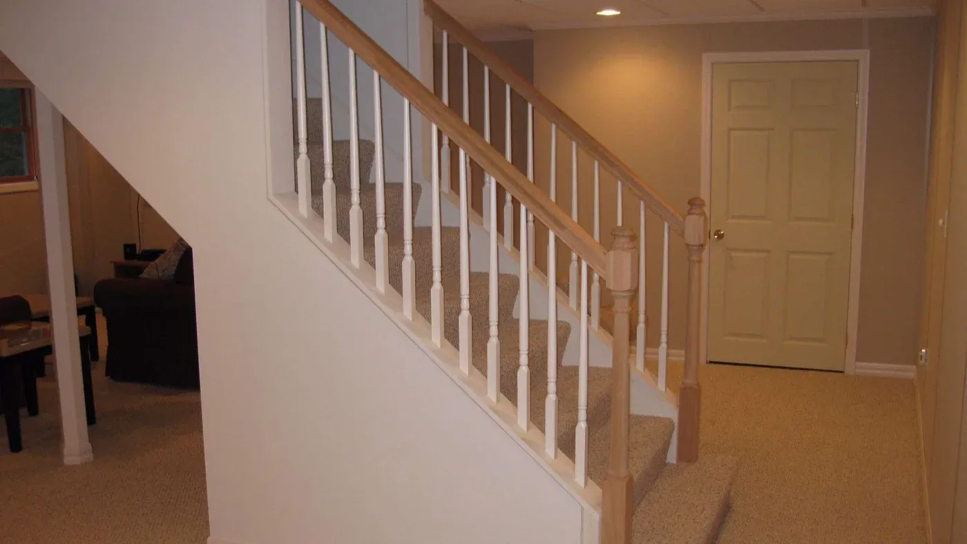 Carpeted staircase with wooden railing in a beige room with soft lighting, leading to a closed door. Sofa visible in the dimly lit adjacent room.