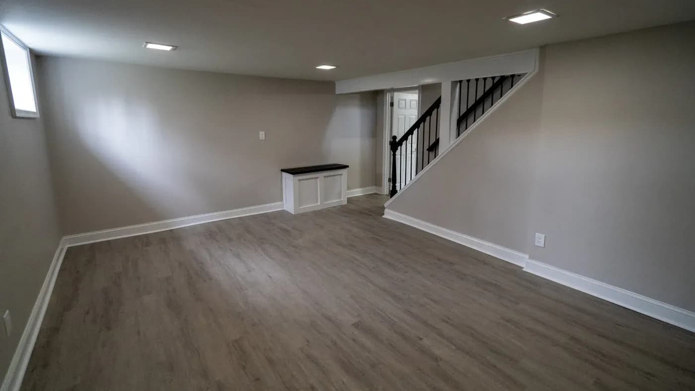 An empty, spacious basement with light gray walls and wood-look flooring. A narrow window lets in light, and a staircase with a black railing is visible.