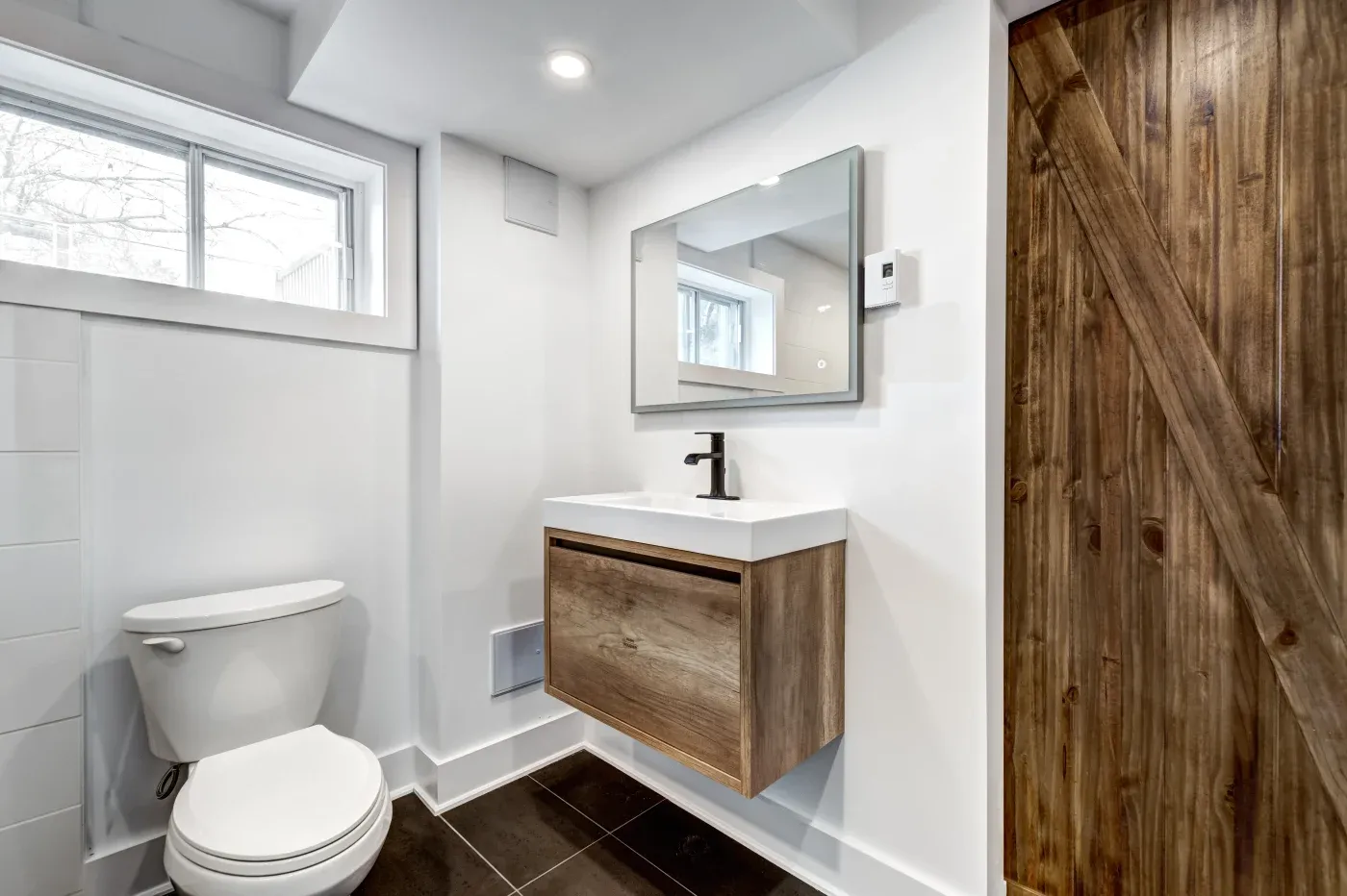 A modern bathroom with white walls, featuring a wooden floating sink with a black faucet under a rectangular mirror. A rustic wooden barn door and toilet are visible, set against a polished, minimalistic design.