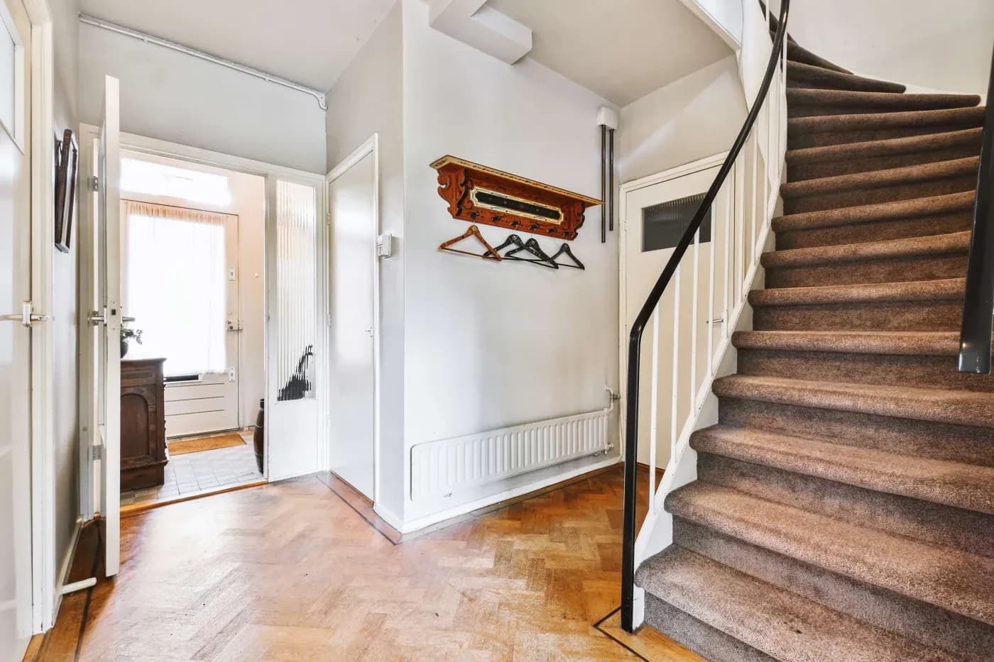 A bright hallway with wooden herringbone flooring, a carpeted staircase on the right, coat rack with hangers, and doors leading to other rooms.