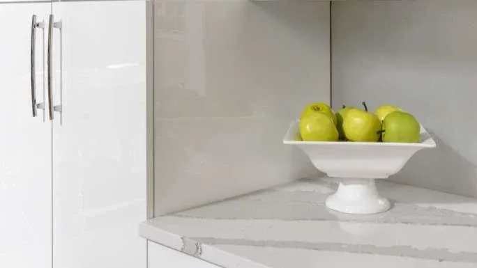 A minimalist kitchen features glossy white cabinets with sleek metal handles. A white pedestal bowl filled with green apples sits on a marble countertop.