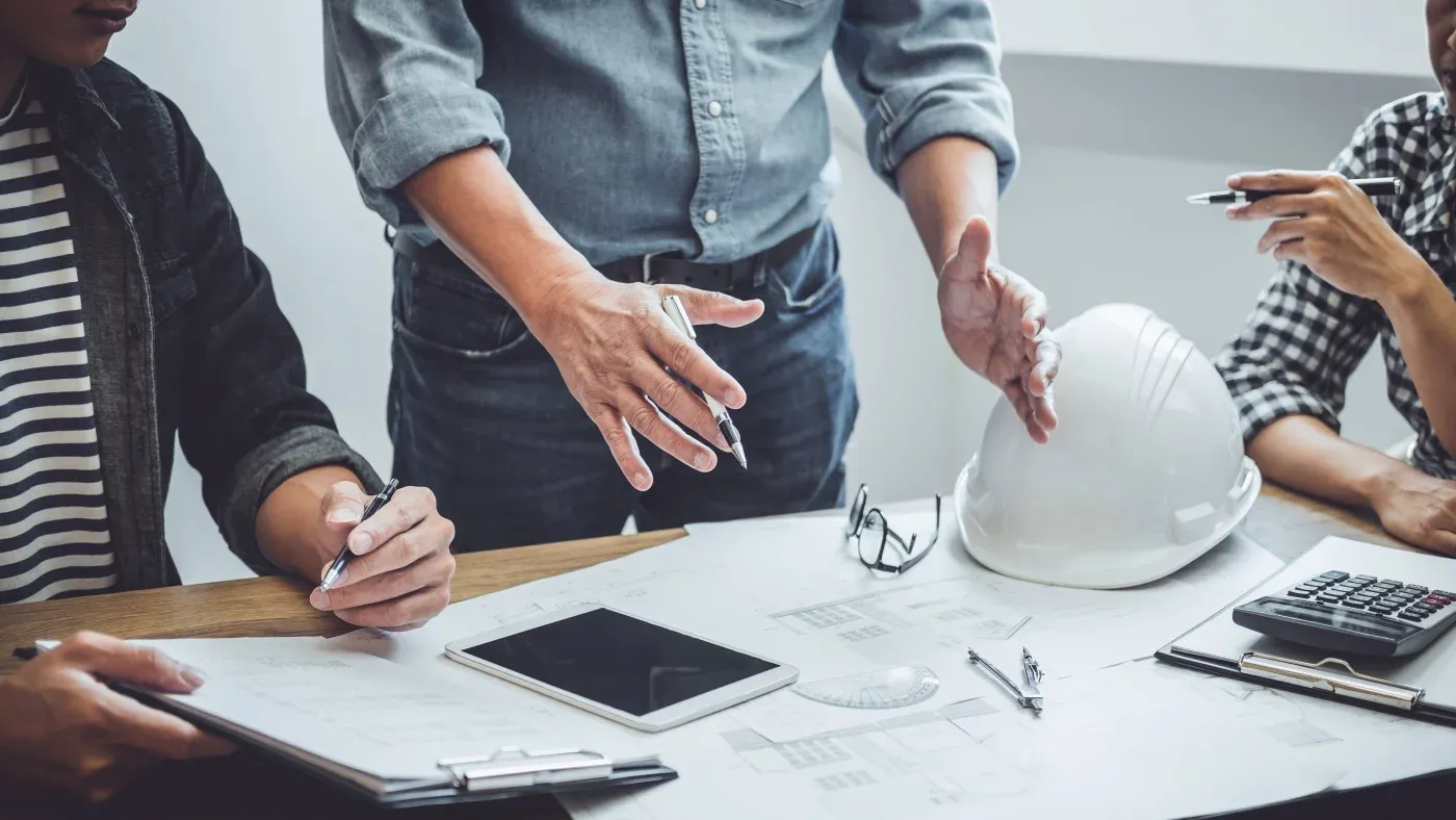 Three people in a meeting discuss architectural plans. A person in the center gestures over blueprints, surrounded by a tablet, hard hat, and calculator.