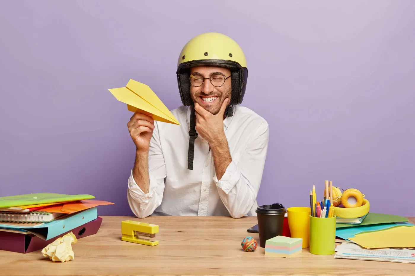 A man wearing a yellow helmet sits at a cluttered desk, smiling while holding a paper airplane. The setting is playful, with colorful office supplies.