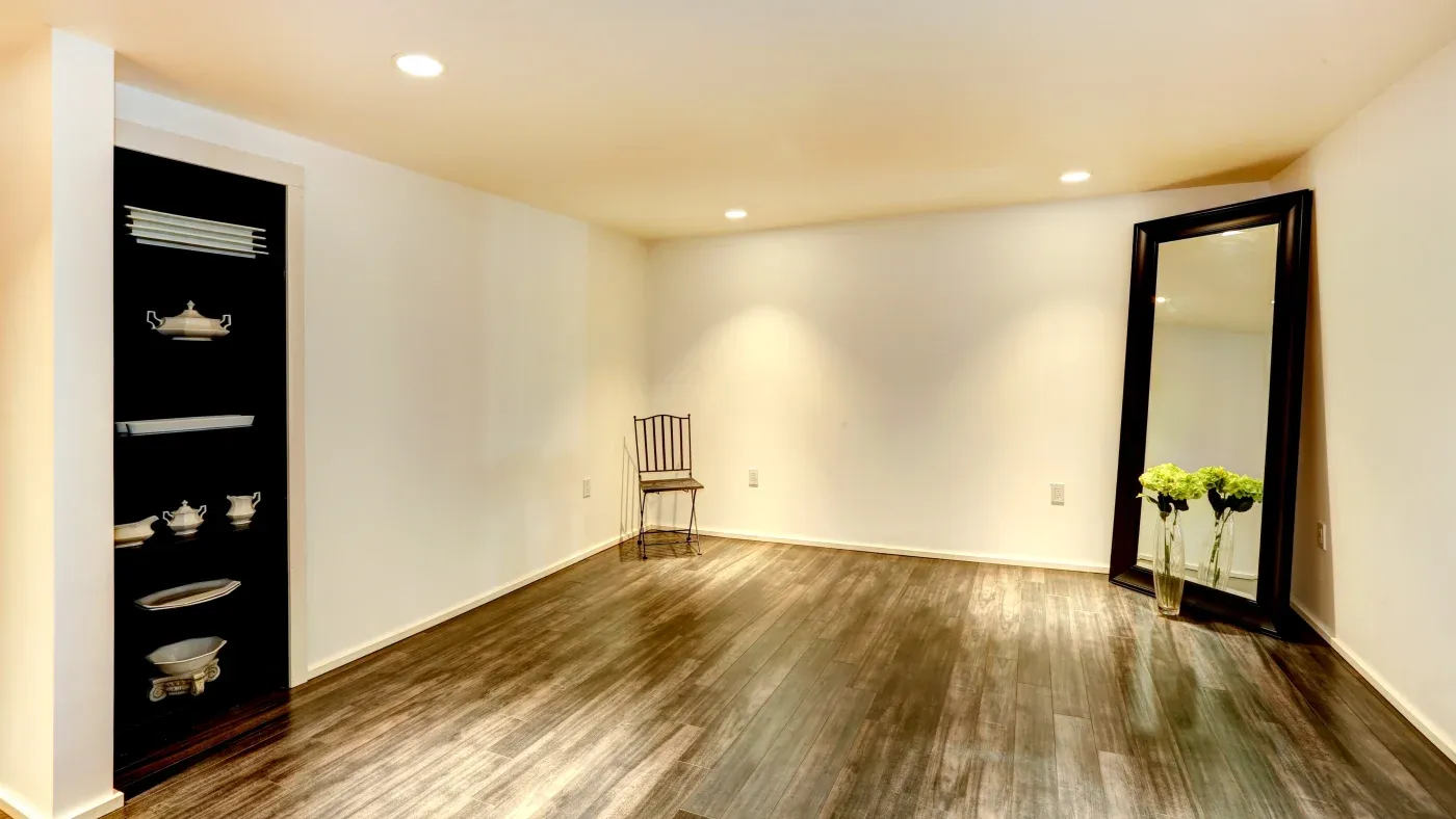 A minimalist room with dark wooden floors, white walls, a large black-framed mirror on the right, a metal chair, and built-in shelves displaying silverware.