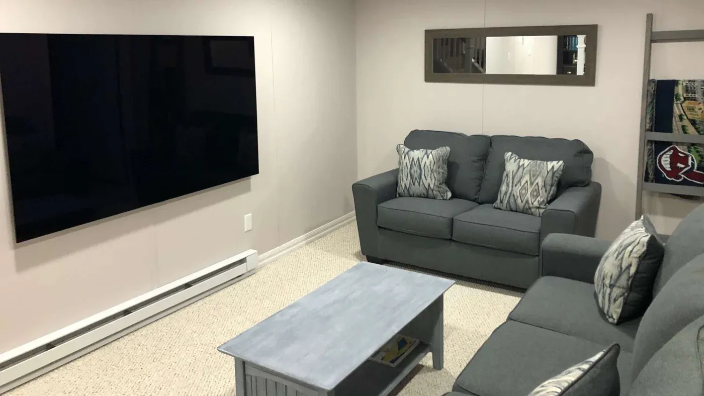 A cozy living room with a large wall-mounted TV, two gray sofas adorned with patterned cushions, and a wooden coffee table. The space feels modern and inviting.