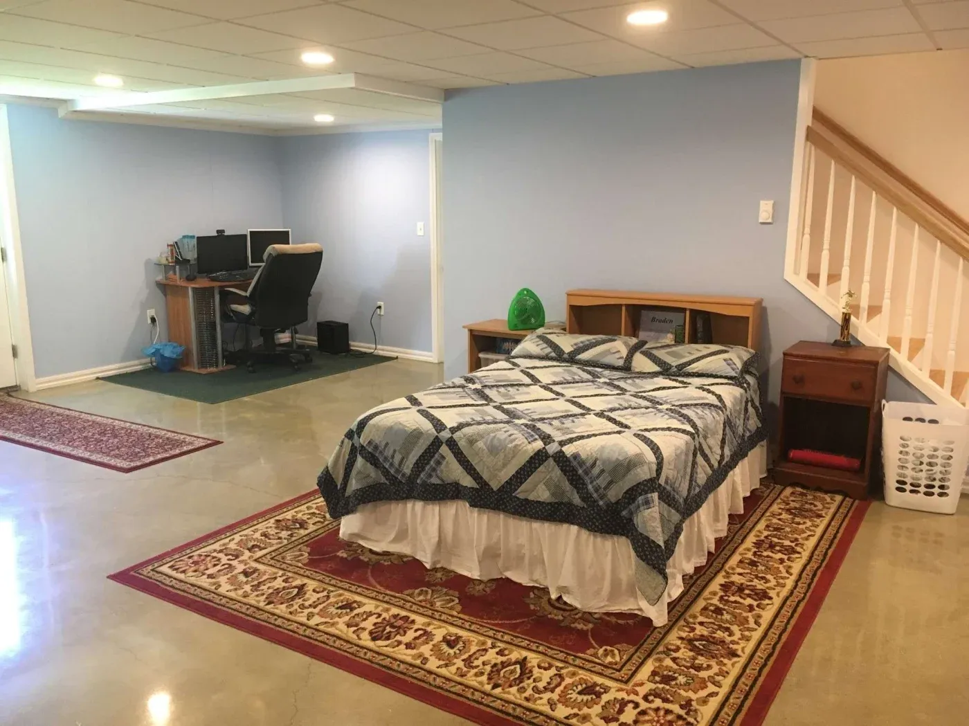 A basement room features a bed with a blue quilt on a patterned rug, a nightstand with a lamp, and a small desk with a computer. The space is well-lit and tidy.