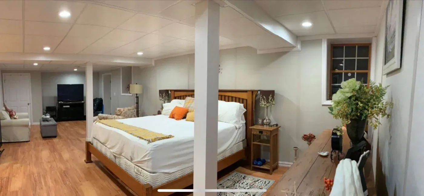 Spacious basement bedroom with a large bed adorned with white and orange pillows. Wooden floors, neutral walls, soft lighting, and a seating area create a cozy, inviting atmosphere.