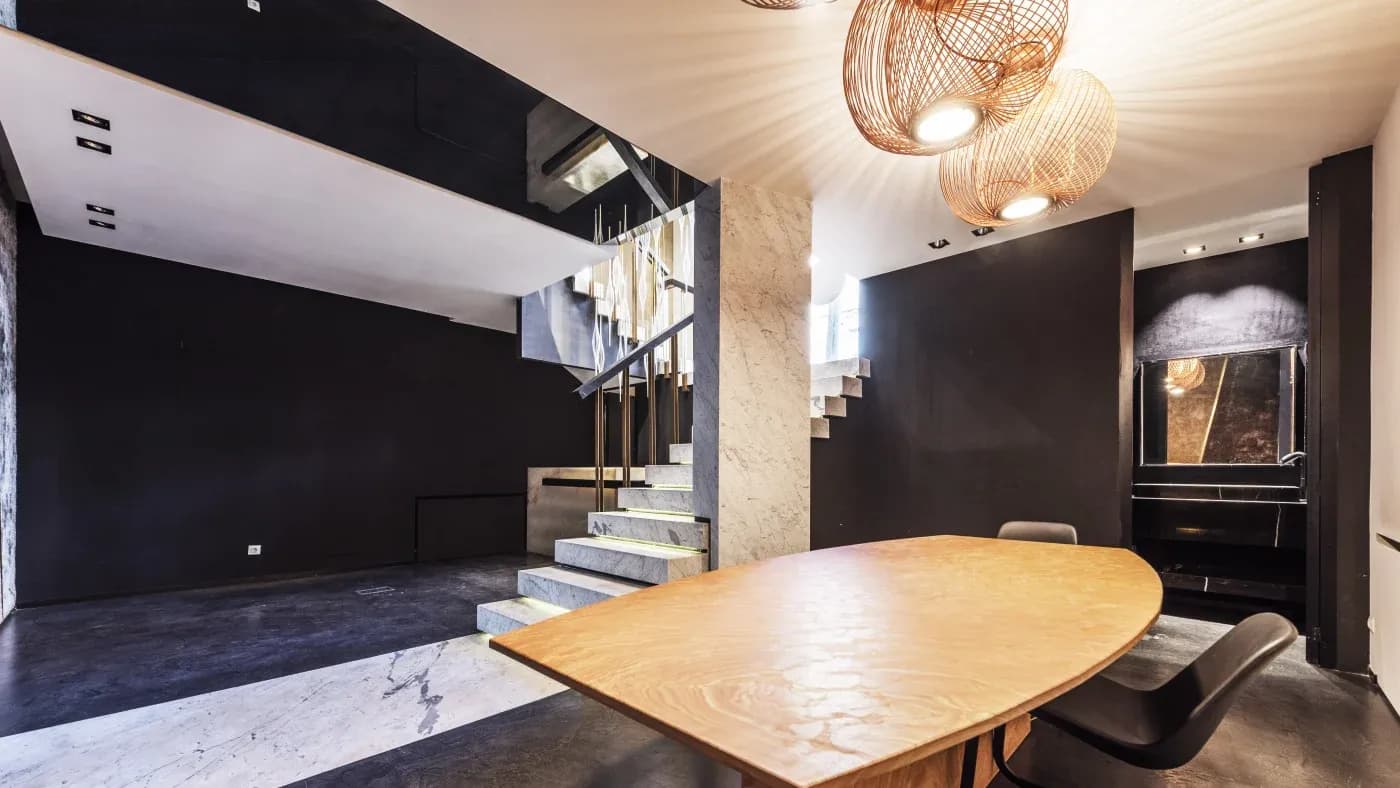 Modern interior with sleek black walls and a floating staircase. A wooden table with black chairs is illuminated by artistic woven pendant lights, creating a stylish, contemporary ambiance.