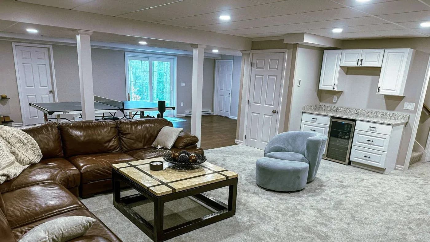 A cozy basement features a large brown leather sectional, a sleek coffee table with decor, a kitchenette, a soft blue chair, and a ping-pong table, creating a relaxed atmosphere.