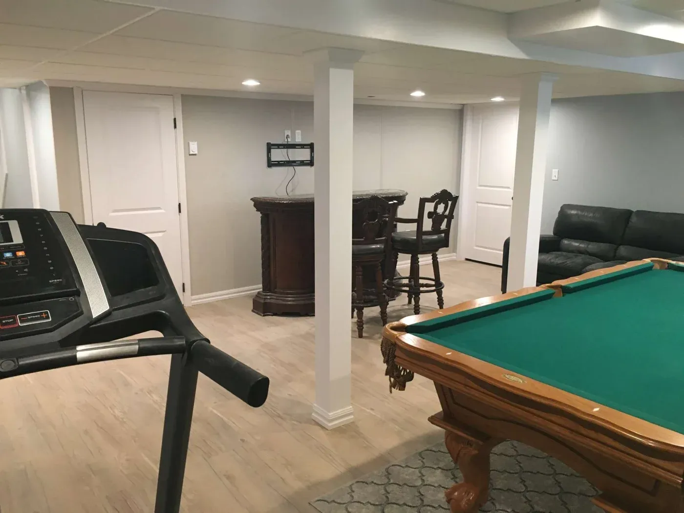 Basement game room with a treadmill, a green pool table, a small bar with stools, and a black leather sofa. The space is cozy and well-lit.