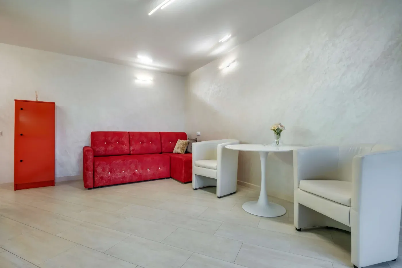 Minimalistic room with a red sofa and cabinet on the left. On the right, a round table with two white chairs and a small vase of flowers.