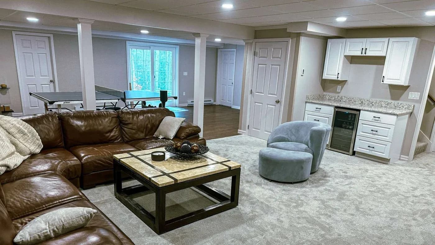 A cozy basement living area features a brown leather sectional, modern coffee table, and blue swivel chair. A kitchenette is in the background, along with a ping pong table near glass doors.