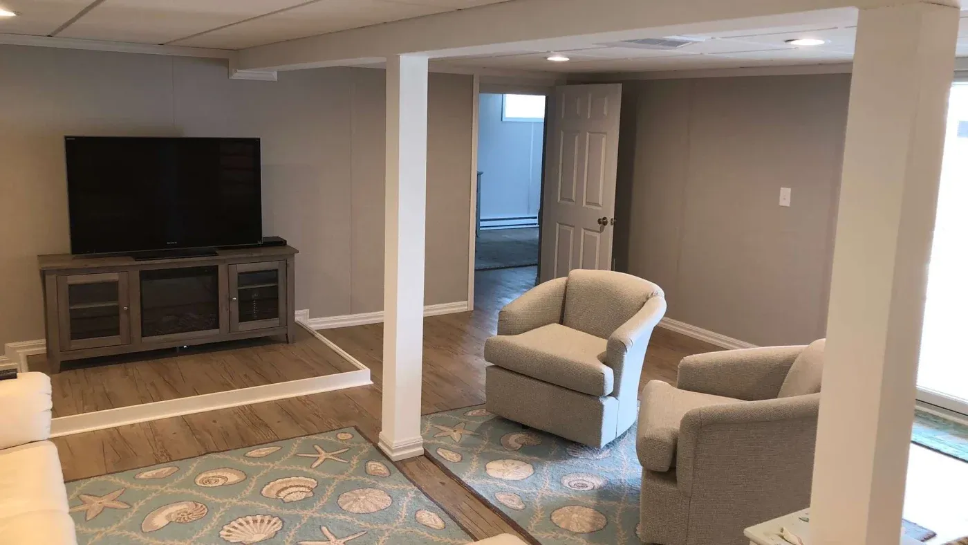 A cozy living room with beige walls features a flat-screen TV on a wooden stand, two upholstered armchairs, and a rug with seashell and starfish designs.