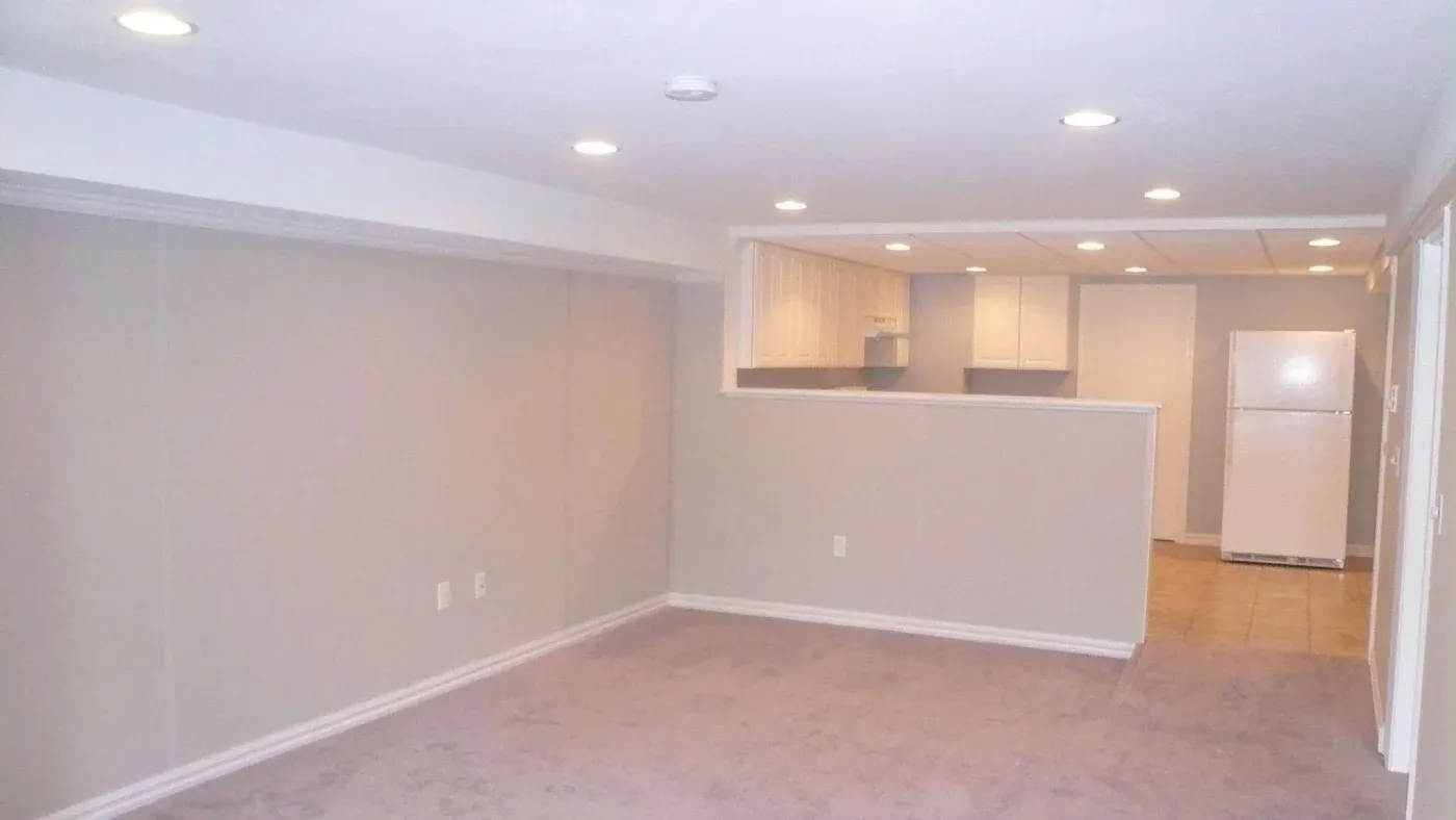 A mostly empty basement room with beige walls and carpet, recessed lighting, and a small open kitchen featuring white cabinets and a refrigerator.