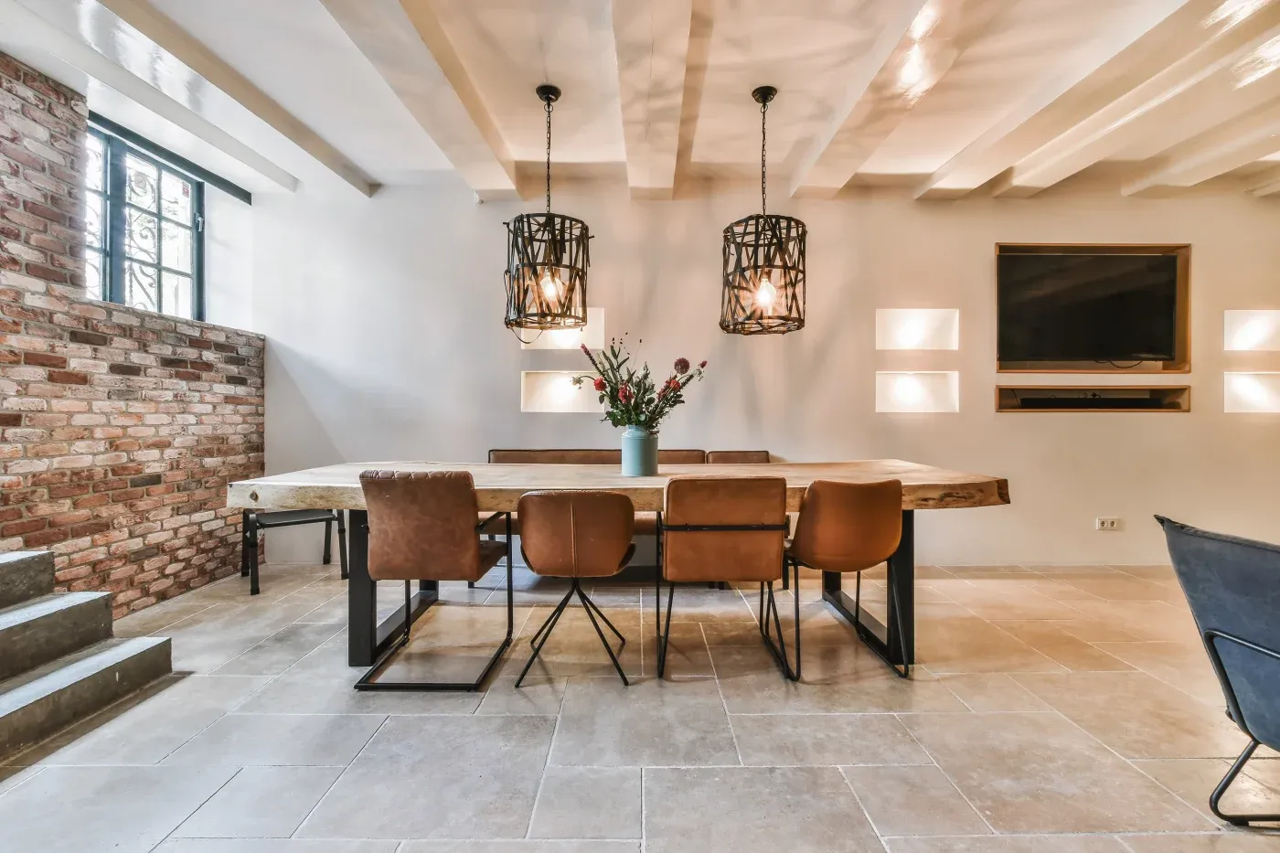 Modern dining room with brick wall and sleek tile floor. A wooden table with six brown chairs is lit by two pendant lamps above, creating a cozy ambiance.
