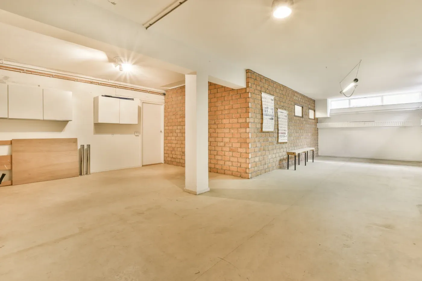 Spacious basement with beige concrete floor and exposed brick wall. Minimalist vibe features white cabinets, wooden panels, and bright lighting.