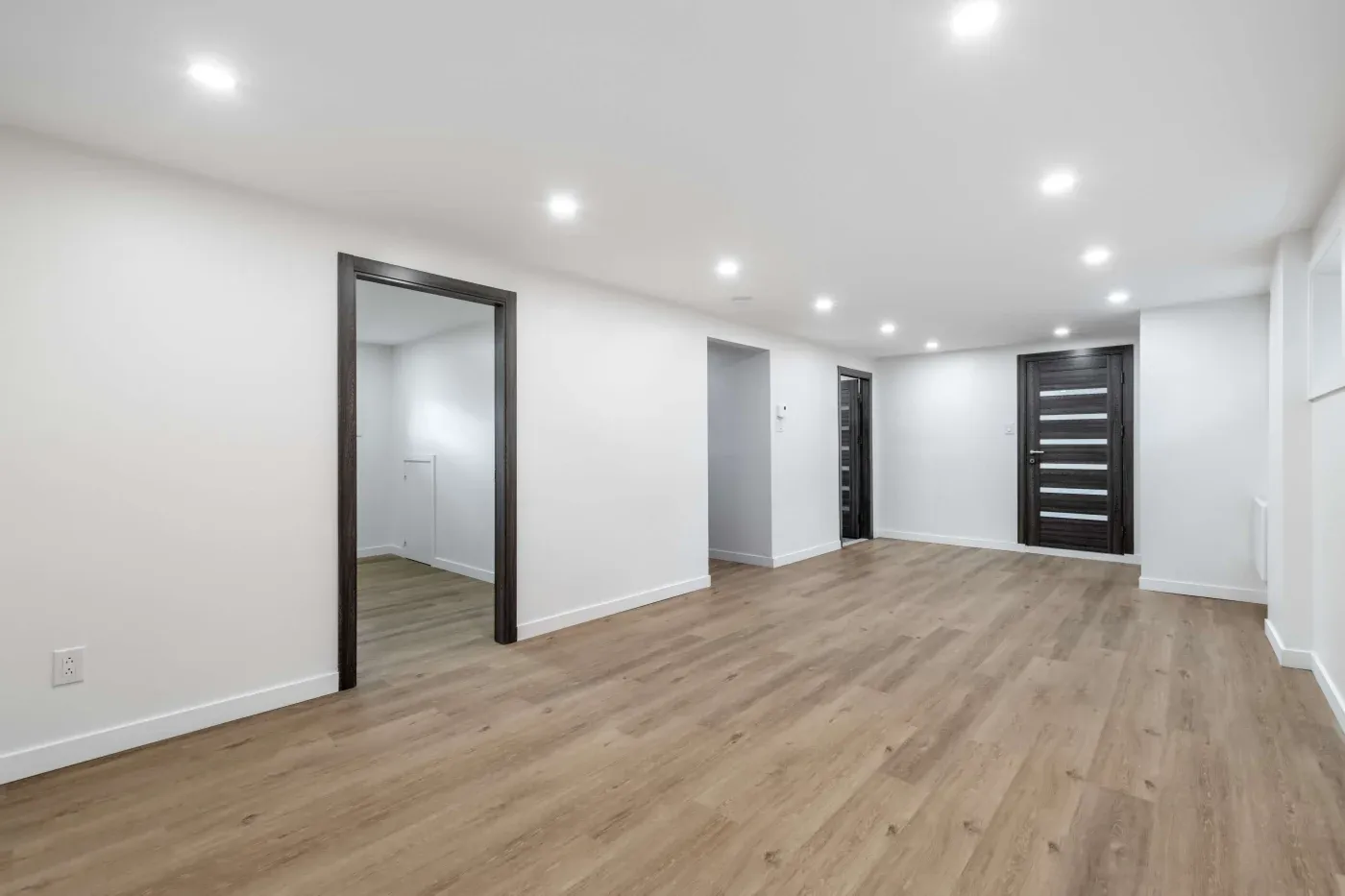 Spacious, modern interior with light wood flooring, white walls, and recessed ceiling lights. Two dark wooden doors add contrast and elegance.