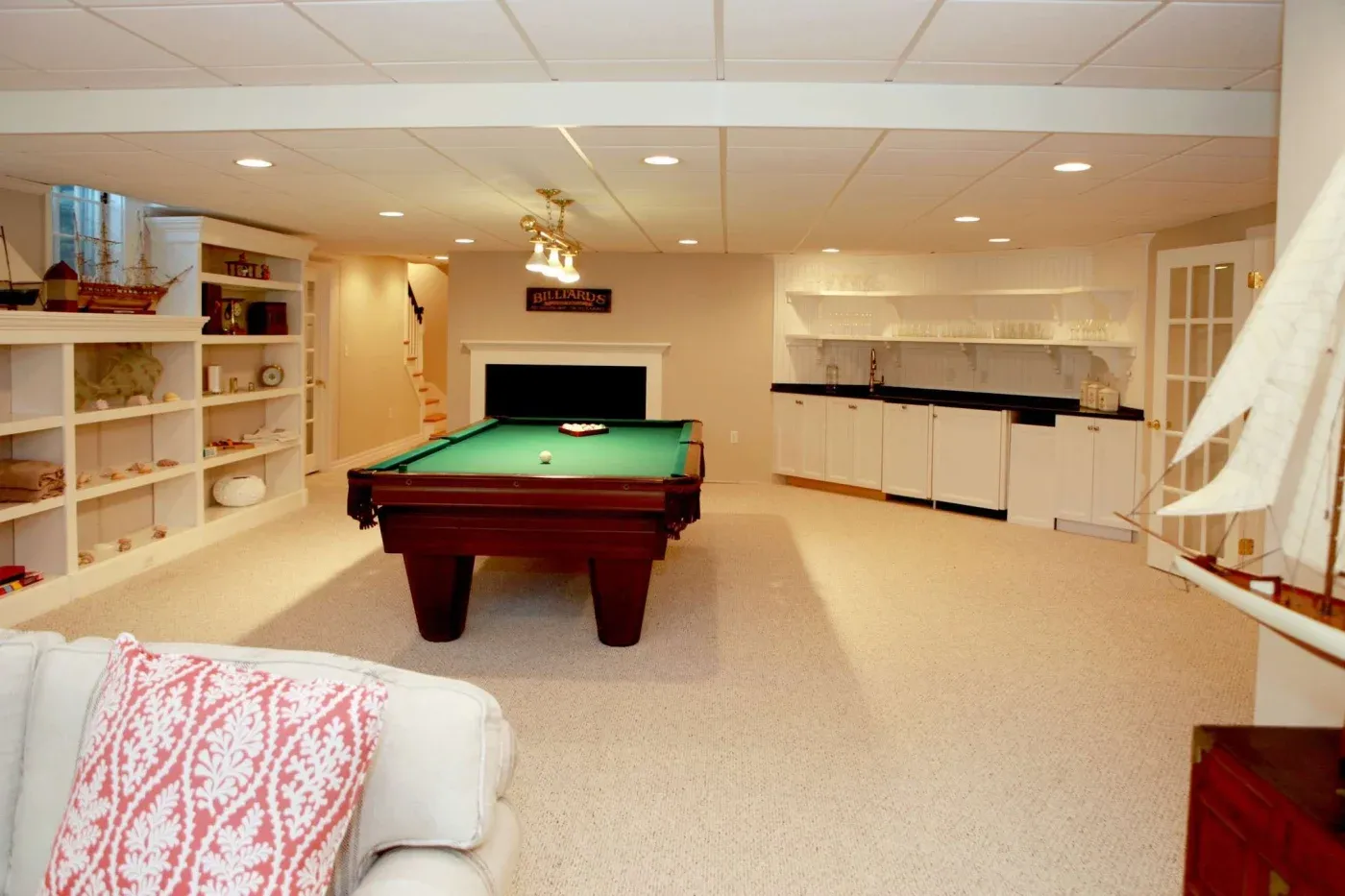 Spacious basement game room with a pool table centered on beige carpet. Shelves with nautical decor and a model ship create a cozy, relaxed atmosphere.