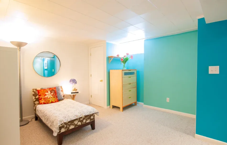 Bright basement bedroom with turquoise walls, a single bed with floral bedding, floral accent pillows, a small dresser with flowers, and a round mirror.