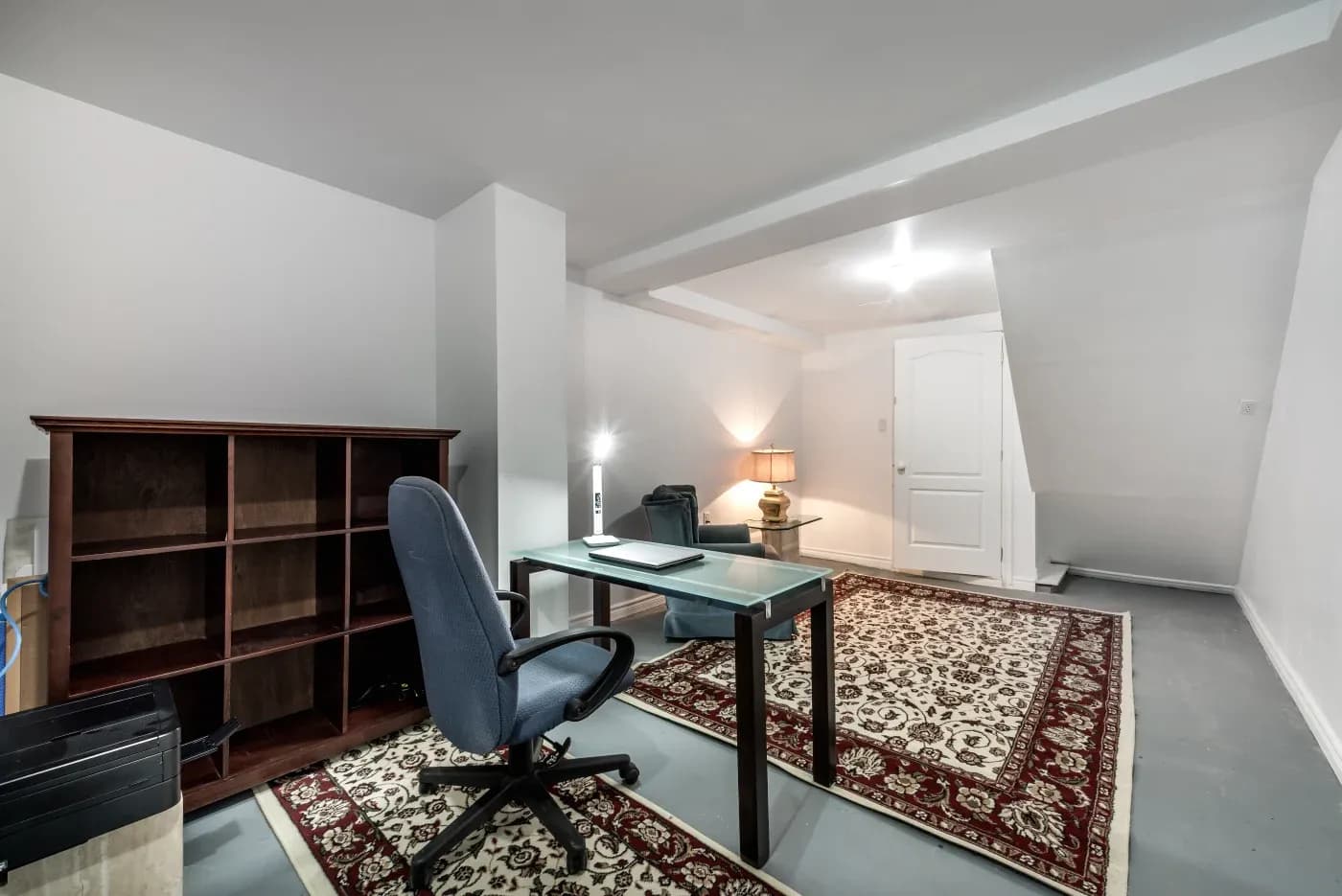 A modern basement office with a glass desk, swivel chair, and laptop. Bookshelf on the left; warm lamp lighting and patterned rugs create a cozy ambiance.