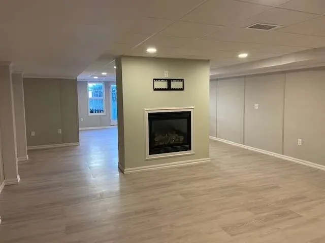 Spacious finished basement with light wood flooring, a central modern fireplace, recessed lighting, and neutral-toned walls, conveying a cozy atmosphere.