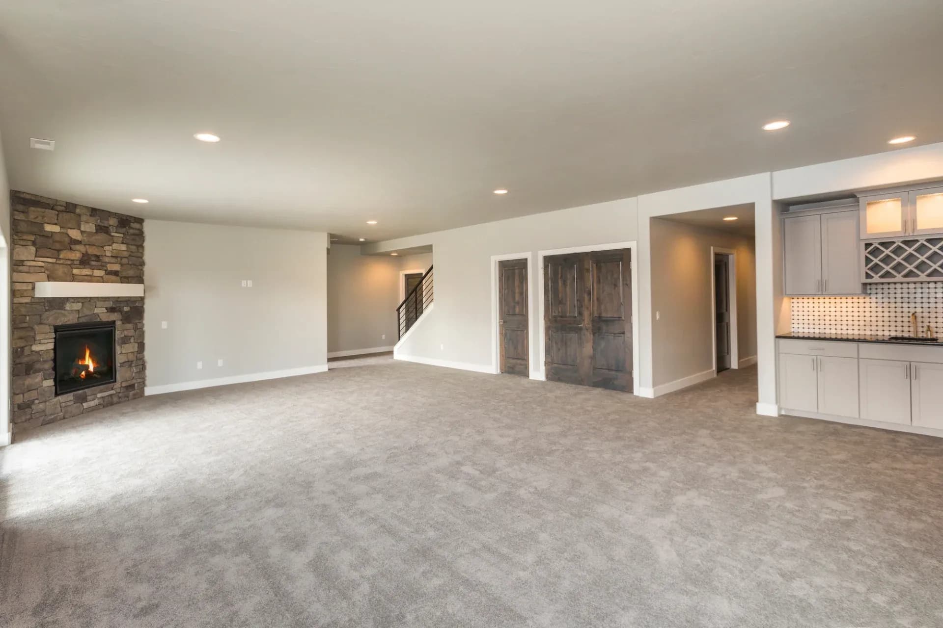 Spacious living area with gray carpet, stone fireplace on the left, and recessed lighting. Wooden doors and a kitchen area with cabinets visible on the right.
